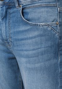 Jasnoniebieskie dżinsy o gładkiej fakturze, z kieszenią z przodu i miedzianymi nitami, prezentujące delikatne przetarcia w tkaninie.