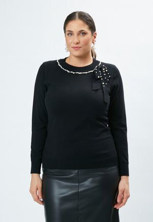 Pullover nero a maniche lunghe con colletto adornato di perle e dettaglio a fiocco laterale. Realizzato in tessuto morbido, silhouette aderente, abbinato a una gonna di pelle.