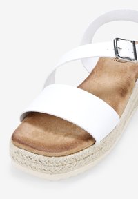 Sandale espadrille blanche avec deux larges brides, une semelle intérieure douce couleur tan et une semelle en jute en forme de coin. Accent en boucle argentée sur la bride.
