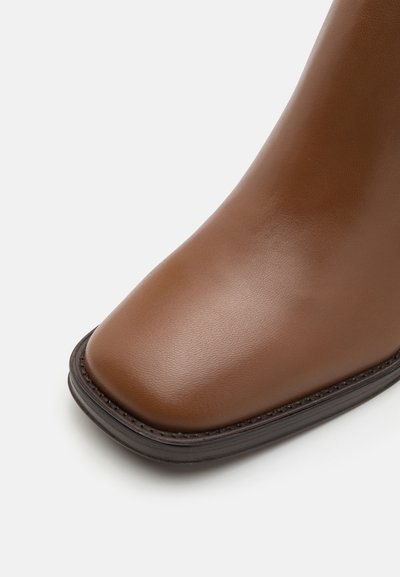 Botte chelsea en cuir marron avec un bout arrondi, texture lisse et semelle en caoutchouc foncé, présentant des coutures discrètes le long du bord.