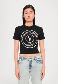 Juodas trumpas marškinėliai su apvaliu logotipu, kuriame užrašyta "Versace Jeans Couture" šviesiai rožine spalva. Trumpomis rankovėmis ir apvaliu iškirpimu.