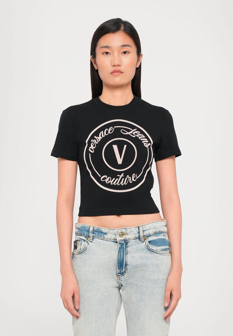 Juodas trumpas marškinėliai su apvaliu logotipu, kuriame užrašyta "Versace Jeans Couture" šviesiai rožine spalva. Trumpomis rankovėmis ir apvaliu iškirpimu.