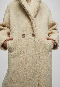 Manteau en polaire de couleur crème avec une surface texturée, un large col châle surdimensionné, une fermeture à double bouton et des poches latérales.