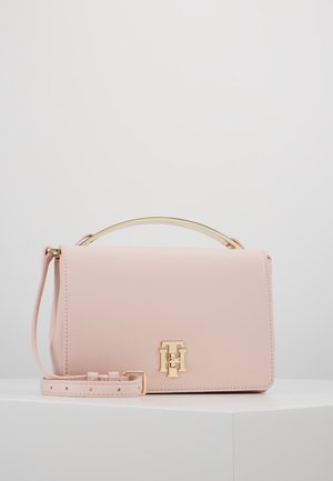 Mała blado różowa skórzana torebka z odpinanym paskiem, złotą metalową rączką i złotym logo TH na przodzie, umieszczona na białej powierzchni.