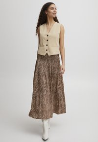 Beige mouwloze vest met een V-hals, zwarte knopen, gecombineerd met een lange, vloeiende rok met een bruine luipaardprint en witte hakken.
