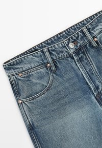 Jean en denim bleu clair avec un design à cinq poches, coutures contrastées, fermeture par bouton argenté et légère décoloration près de la cuisse.