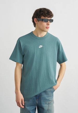 Ung mann iført teal Nike t-skjorte, blå jeans, svarte rektangulære solbriller, stående med en hånd i lomma mot ensfarget bakgrunn.