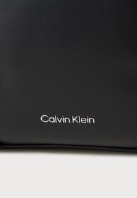 Borsa nera in pelle sintetica con una texture liscia. Presenta il logo "Calvin Klein" in lettere metalliche argentate nella parte inferiore.