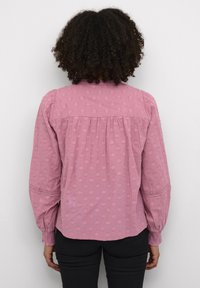 Blouse rose en tissu texturé avec des motifs en relief, dos froncé et poignets de manches élastiques, avec une délicate bordure en dentelle.