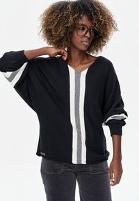 Zwarte oversized trui met witte en grijze verticale strepen, V-hals ontwerp en opgerolde manchetten. Gemaakt van zacht, gestructureerd breimateriaal.