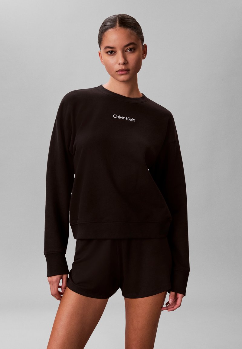 Γυναίκα με μαύρο μακρυμάνικο φούτερ Calvin Klein και μαύρο σορτς που ταιριάζουν, στέκεται μπροστά σε απλό γκρι φόντο.