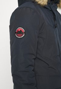 Granatowa puchowa kurtka z kapturem wyłożonym futrem, z widocznym czerwono-czarnym haftowanym logo na lewym rękawie.