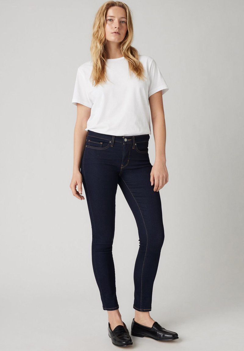 Jeans ajustados azul oscuro con costuras en contraste, combinados con una camiseta blanca sencilla y mocasines negros, sobre un fondo neutro.