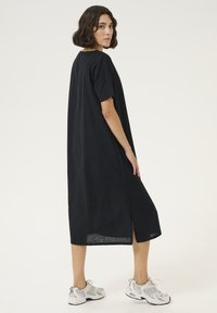 Schwarzes knielanges Kleid aus leichtem Stoff mit kurzen Ärmeln, seitlichen Schlitzen und lockerer Passform, getragen mit grauen Sneakers.