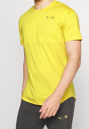 Muž oblečený v jasně žlutém tričku s krátkým rukávem značky Puma a tmavých šortkách s logy Puma a "First Mile" na obou kusech oblečení.