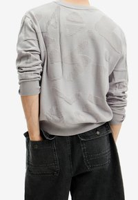 Szary sweter z teksturowanymi abstrakcyjnymi wzorami, ściągaczami na mankietach i okrągłym dekoltem, zestawiony z ciemnymi dżinsami z dużymi kieszeniami.