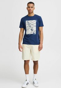 Marineblauw T-shirt met een grafisch ontwerp dat cijfers en lijnen bevat, gecombineerd met lichtbeige shorts. Het shirt heeft een ronde hals en korte mouwen.