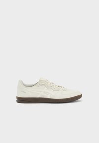 Bílá kožená teniska s nízkým profilem, obsahuje béžové akcenty, strukturované boky a hnědou gumovou podrážku. Obsahuje také perforované detaily.
