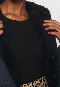 Svart ribbad topp under en marinblå fluffig jacka, med stora knappar och en mjuk textur. Leoprintdetaljer på nederkanten.