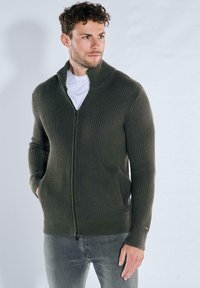 Olive green ribbed sweater featuring a high neck, zipper closure, and side pockets, made of textured knit fabric.