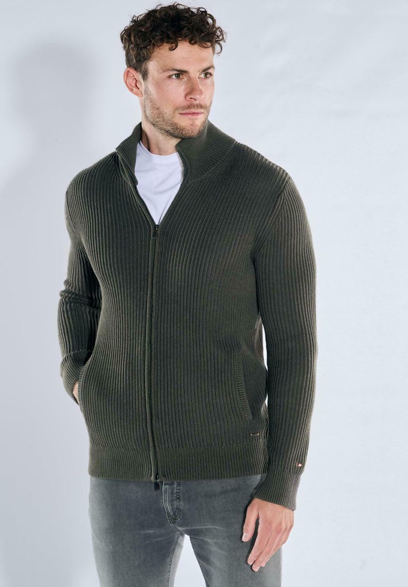 Olive green ribbed sweater featuring a high neck, zipper closure, and side pockets, made of textured knit fabric.