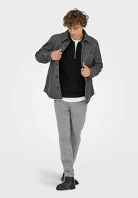 Grau karierter Blazer über einem schwarzen Pullover, kombiniert mit grauen Hosen und schwarzen Stiefeletten. Der Blazer hat eine knöpfbare Front und eine Brusttasche.
