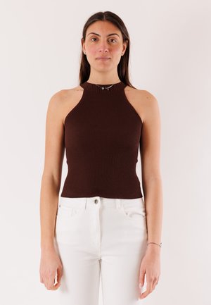 Donna con capelli lunghi e scuri che indossa una canottiera marrone a costine e pantaloni bianchi, rivolta in avanti contro uno sfondo chiaro e uniforme.