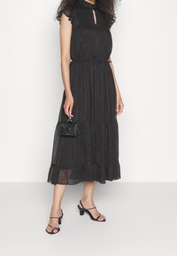 Robe maxi noire avec un col à volants, un décolleté en trou de serrure et un ourlet à volants superposés. Associée à un petit sac à main noir et des talons à lanières.