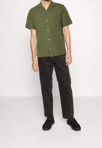 Camicia verde oliva a maniche corte con tasca sul petto, chiusura a bottoni, abbinata a pantaloni scuri e scarpe nere. Design semplice e casual.