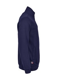 Navy sininen vetoketjuhuppari, joka on valmistettu pehmeästä kankaasta. Ominaisuuksiin kuuluvat korkea kaulus, sivutaskut, ribbireunukset ja pieni logo helmoissa.