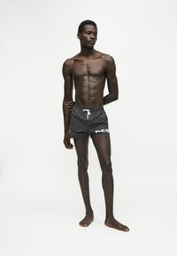 Homme mince à la peau foncée debout pieds nus portant un short de bain noir à motifs avec un cordon blanc sur un fond clair uni.