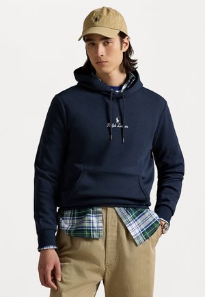 Marineblauer Hoodie mit Kängurutasche, mit "Ralph Lauren" Stickerei. Über einem karierten Hemd getragen, kombiniert mit khakifarbenen Hosen und einer beigen Mütze.