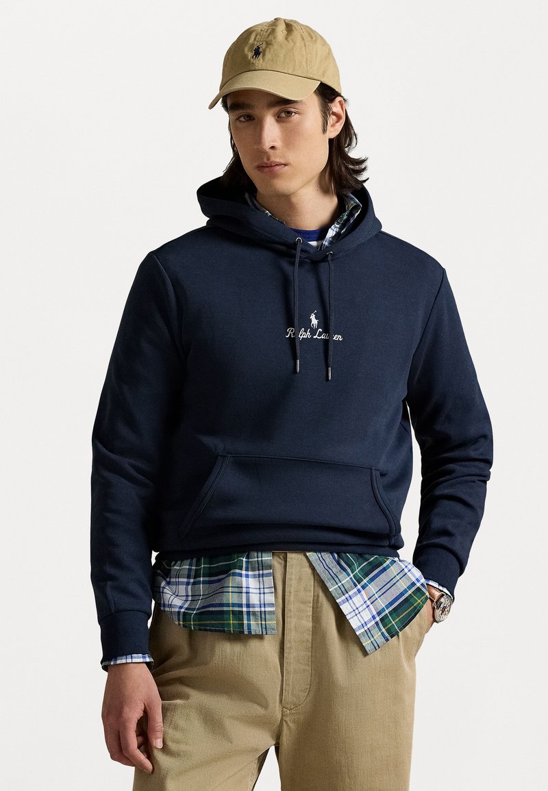 Jūras zila kapuce ar kangarū pirkstu kabatu, ar "Ralph Lauren" izšuvumu. Kombinēta ar flaneļa kreklu, saskaņota ar khaki biksēm un bēša cepuri.