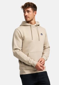 Beige hoodie gemaakt van zacht materiaal met verstelbare trekkoord, een voorkangoeroezak en een klein rond logo op de borst.