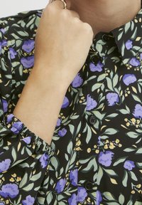 Svart blommig blus med lila blommor och gröna blad, med elastiska manschettdetaljer och svarta knappar framtill.