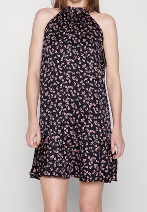Femme portant une robe noire sans manches à motif floral rose et blanc avec un ourlet à volants, debout devant un fond clair uni.