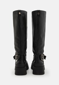 Botas negras hasta la rodilla con un exterior de cuero suave, suelas de goma gruesa, detalles de hebilla y una costura prominente en la parte posterior.