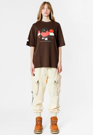 Camiseta oversized marrón con estampado gráfico, combinada con pantalones cargo beige con bolsillos con cremallera y botas color tostado con cordones.