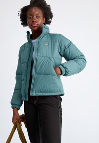 Zeleno modra puhovka z zadrgo, rebrastimi manšeti in vzorčasto tkanino. Model drži svetlo rjav nahrbtnik, v kombinaciji z črnimi hlačami.