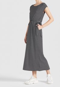 Kvinna i en svart och vit horisontellt randig midiklänning med sidofickor och vita chunky sneakers på en enkel bakgrund.