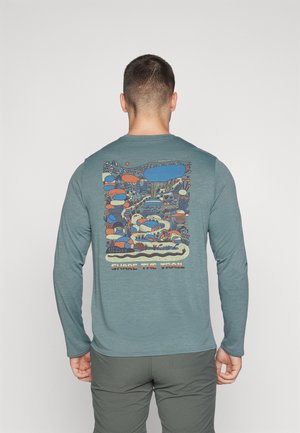 Homme portant une chemise verte à manches longues avec un design abstrait coloré de la nature et le texte « Partagez le sentier » au dos, debout devant un arrière-plan uni.