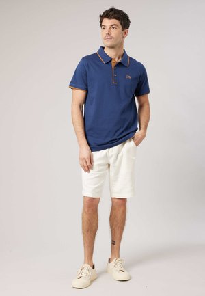 Homme portant un polo bleu avec une bordure marron, un short blanc et des baskets blanches, debout avec une main dans la poche sur un fond neutre.