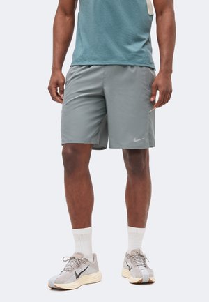 Homme portant un short de sport gris Nike, des chaussures de course beige avec le logo Swoosh noir, des chaussettes blanches et un t-shirt bleu-vert, debout sur fond blanc.