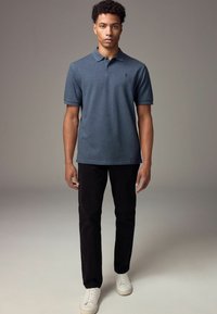 Polo azul oscuro con una textura sutil, mangas cortas y un pequeño detalle bordado, combinado con pantalones negros y zapatillas blancas.