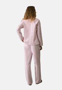 Mujer de pie con la mano en la cadera, vistiendo un conjunto de pijama de manga larga a rayas en rosa claro con pantalones a juego, sobre un fondo liso.