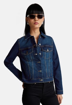 Femme avec des cheveux noirs raides portant une veste en denim bleu foncé, un pantalon noir, des boucles d'oreilles en argent et des lunettes de soleil teintées marron.