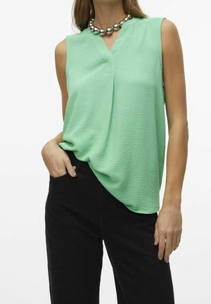 Vrouw draagt een mouwloze lichtgroene blouse met structuur en V-hals en zwarte broek, poseert met één hand in de zak en ketting zichtbaar.