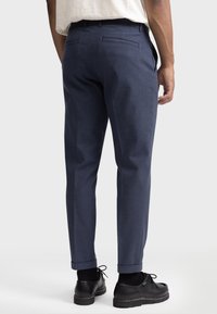 Navyblauwe nauwsluitende broek met een gestructureerde afwerking, omgeslagen zoom en twee achterzakken, gecombineerd met zwarte veter schoenen.