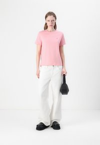 T-shirt di cotone rosa con scollo rotondo, abbinata a jeans bianchi a gamba larga. Il modello tiene una piccola borsa nera; scarpe con tacco spesso nere.