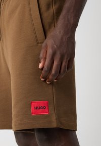 Shorts din bumbac maron cu buzunare laterale. Prezintă o etichetă roșie cu "HUGO" în negru. Prim-plan asupra texturii țesăturii și a fitului.